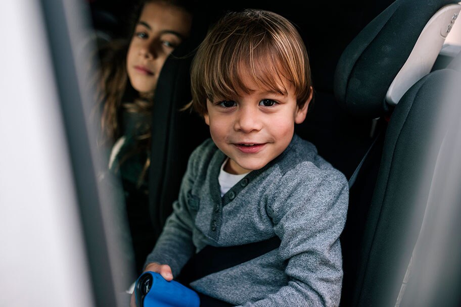 Un niño sonríe mientras se sienta en el asiento trasero del auto abrochado en su asiento para auto. Su hermana mayor está sentada a su lado apareciendo detrás del asiento del auto.