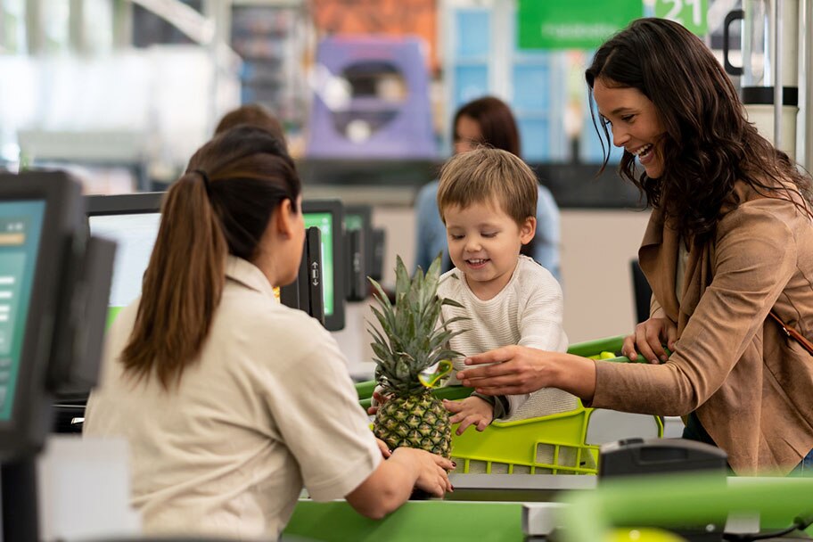 Un niño joven sentado en el carrito de la compra ayuda a su sonriente madre a darle una piña fresca a un cajero en la caja.