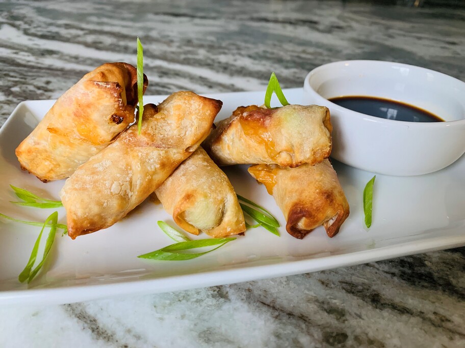 Cuatro rollitos de primavera de pollo a la naranja en un plato con guarnición de cebolla verde y un tazón de salsa para mojar.