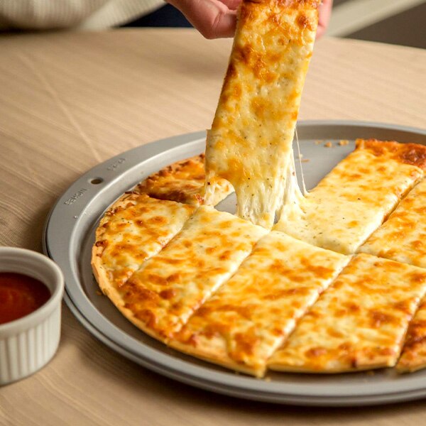 Una mano levanta un Cheesy Pizza Dipper de una bandeja para hornear junto a una taza de marinara.