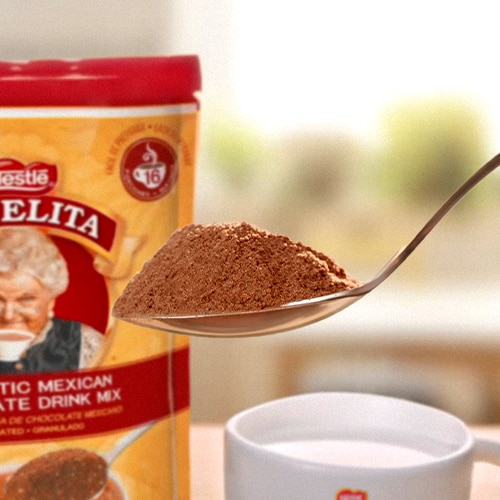 Una cuchara sirve chocolate en polvo directo del empaque de ABUELITA granulado en una taza de leche caliente.