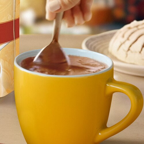 Una cuchara revuelve el chocolate caliente en una taza frente al empaque de chocolate granulado y una concha.
