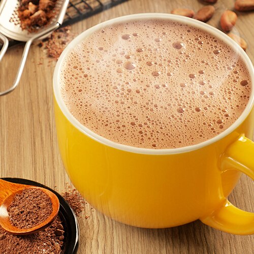 Una taza amarilla de espumoso chocolate caliente junto a cucharas colmadas de chocolate en polvo.
