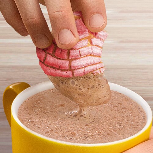 Una mano sopea una concha rosada en una taza amarilla con chocolate.