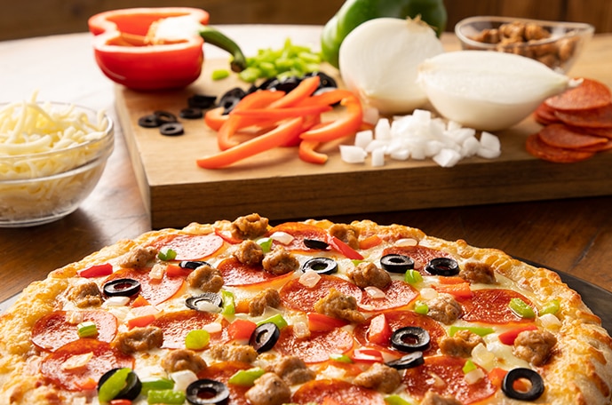 Una pala de madera para pizza y una tabla con pepperoni rebanado, cebollas, pimientos, y hierbas de olor repartidos alrededor de la superficie de madera.