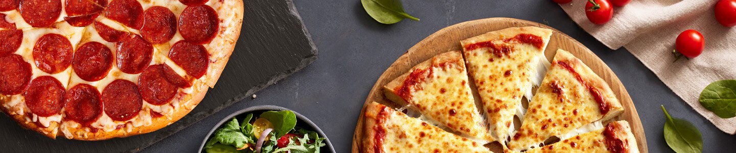 Pizzas de queso y pepperoni sobre tablas de cortar junto a una ensalada, tomatitos y albahaca sobre un fondo gris.