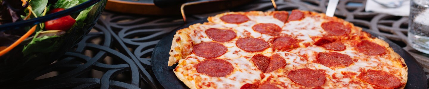 Pepperoni pizza on a black plate near a house salad on a grated table.
