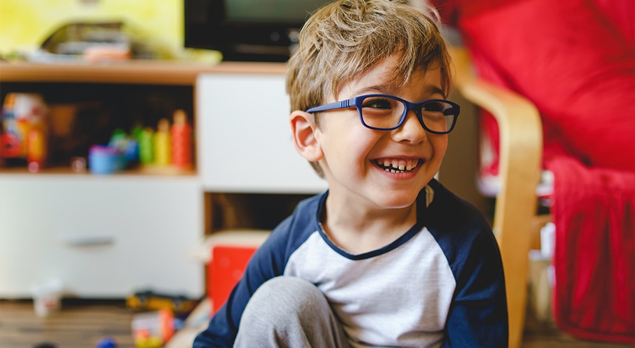 child with glasses smiling on the floor in a living room with toys.