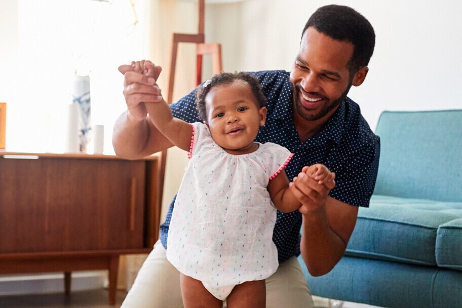 Child smiling with their father who is aiding them in walking upright in a living room.