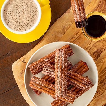 A bowl of churros on a cutting board beside a mug of Abuelita® and a cup of dipping chocolate.