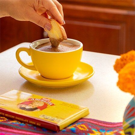 Un empaque de marquetas ABUELITA junto a una taza color amarillo de espumoso chocolate caliente.